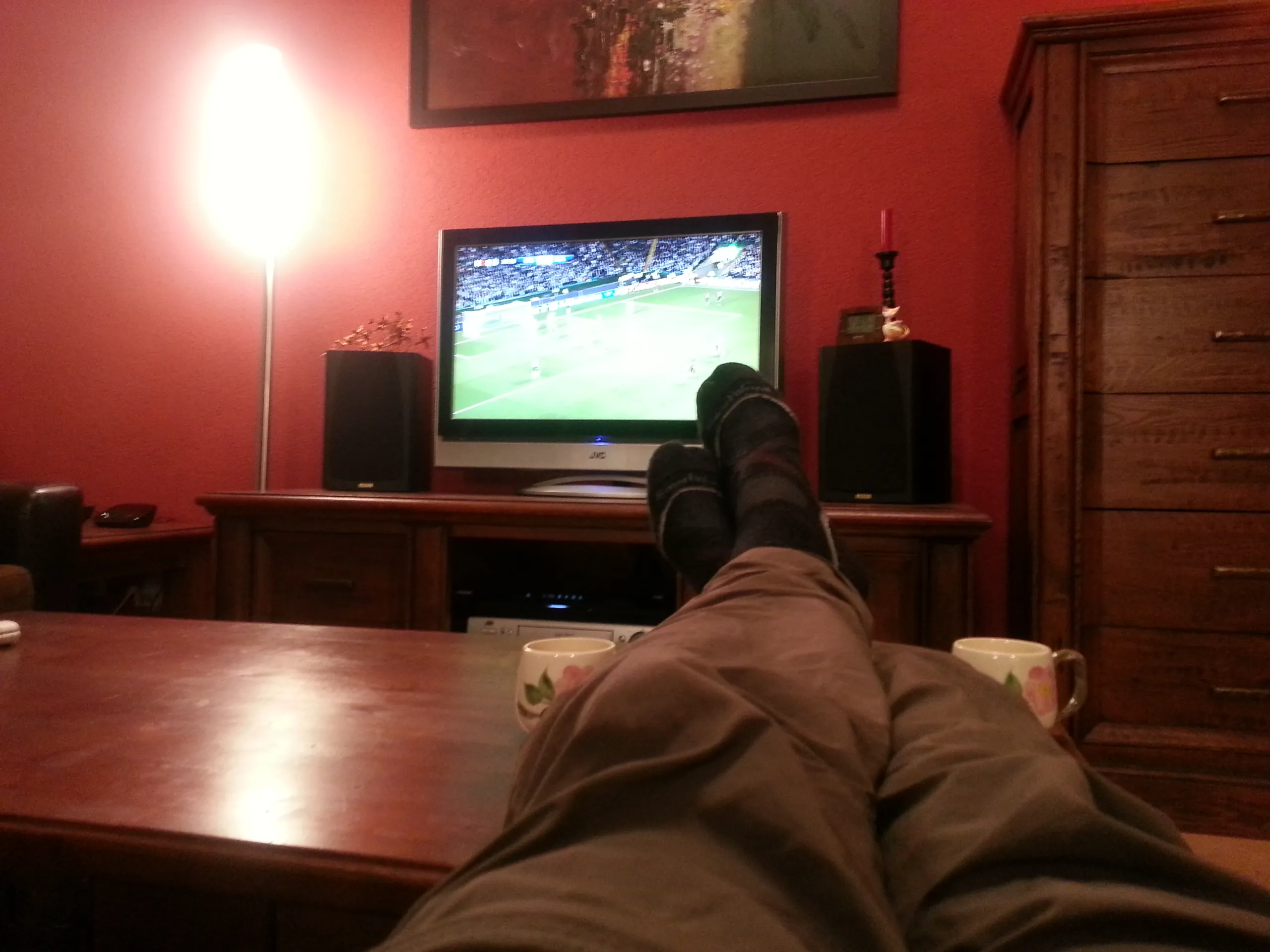 Feet in socks resting on a wooden table in a living room with a red wall, a lamp and a television