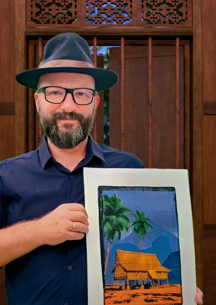 Daniel Arendzen in a hat and glasses holding his gel plate monotype Mystic Mountains, in front of a carved window inside a render of the traditional house he modelled