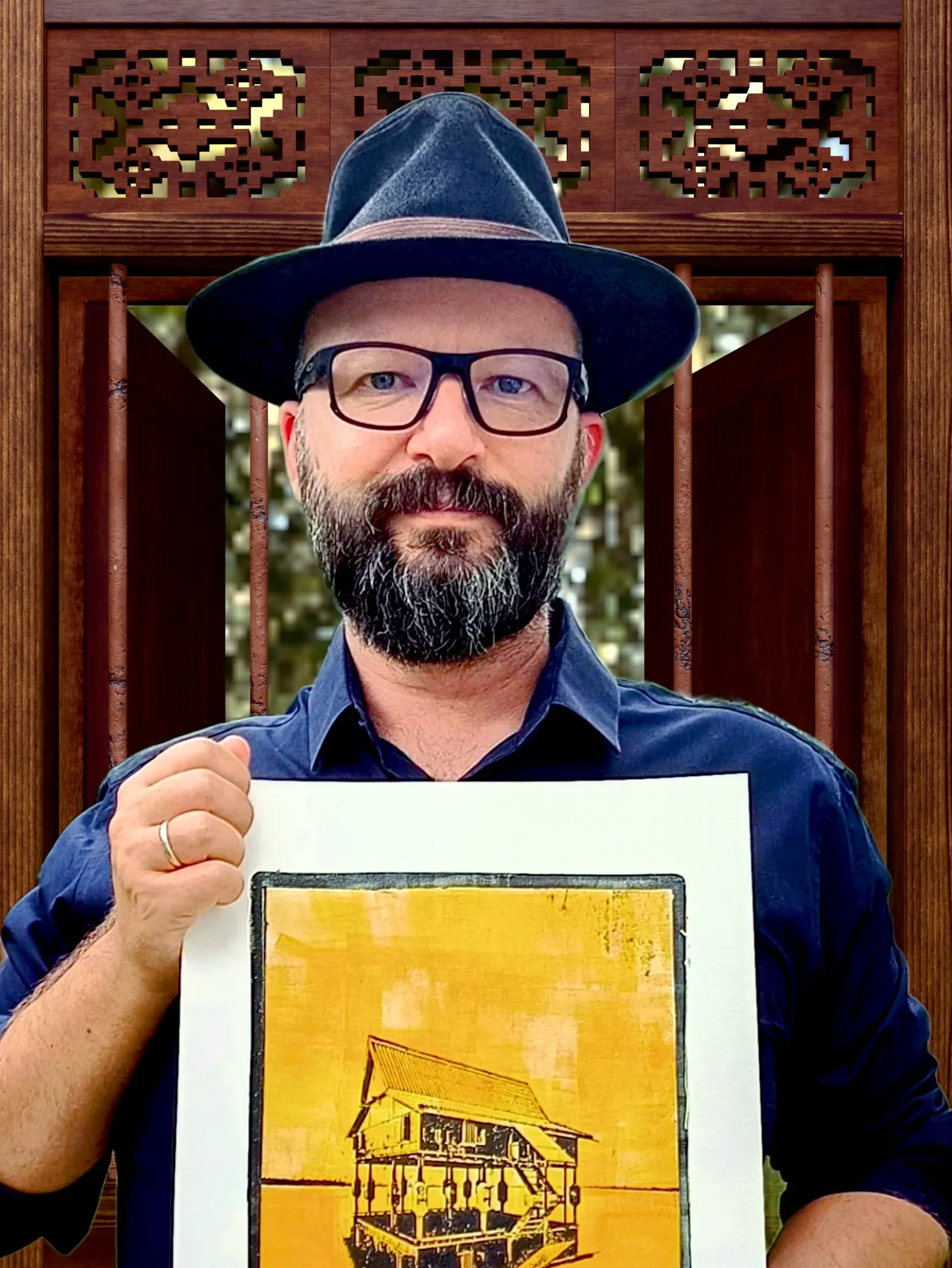 Daniel Arendzen in a hat and glasses holding a yellow gel plate monotype of a traditional Malaysian house, standing inside a Blender render of the same house, with a carved wooden window behind him.