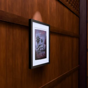 Frame 526 in Purple Shades, gel plate monotype by Daniel Arendzen, framed on a wooden wall under a spotlight, seen from the side, with binoculars, a watch and a lighter on a table below. The room is a Blender render of the interior of the same traditional Malaysian house shown in the print.