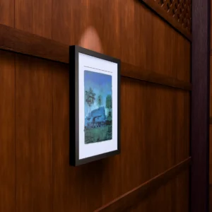 Frame 526 in Winter, gel plate monotype by Daniel Arendzen, framed on a wooden wall under a spotlight, seen from the side, with binoculars, a watch and a lighter on a table below. The room is a Blender render of the interior of the same traditional Malaysian house shown in the print.