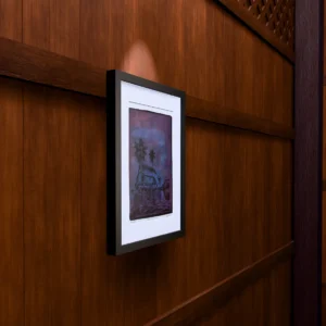 Frame 529 in Purple (l), gel plate monotype by Daniel Arendzen, framed on a wooden wall under a spotlight, seen from the side, with binoculars, a watch and a lighter on a table below. The room is a Blender render of the interior of the same traditional Malaysian house shown in the print.
