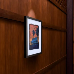 Frame 531 #3 - Wall Frame 531 #3, gel plate monotype by Daniel Arendzen, framed on a wooden wall under a spotlight, seen from the side, with binoculars, a watch and a lighter on a table below. The room is a Blender render of the interior of the same traditional Malaysian house shown in the print.