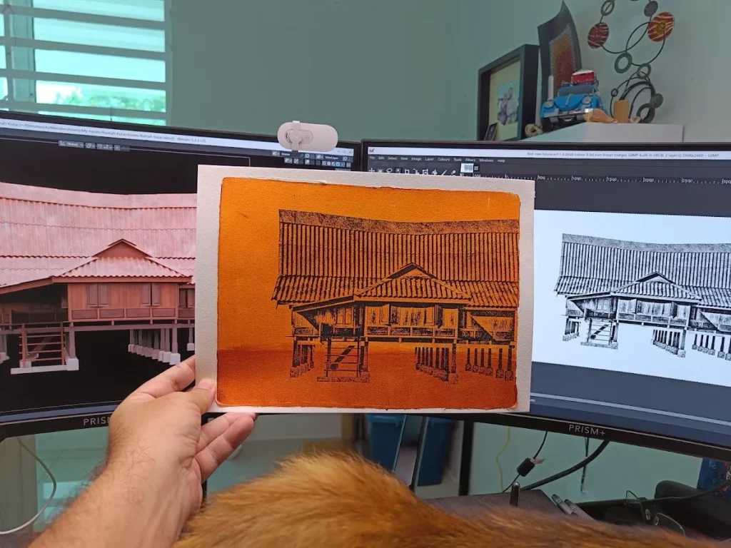 Daniel Arendzen holding a gel plate monotype of a traditional Malaysian house on stilts, in black on orange, in front of his computer screens showing the 3D model of the same house, with a ginger cat in the foreground