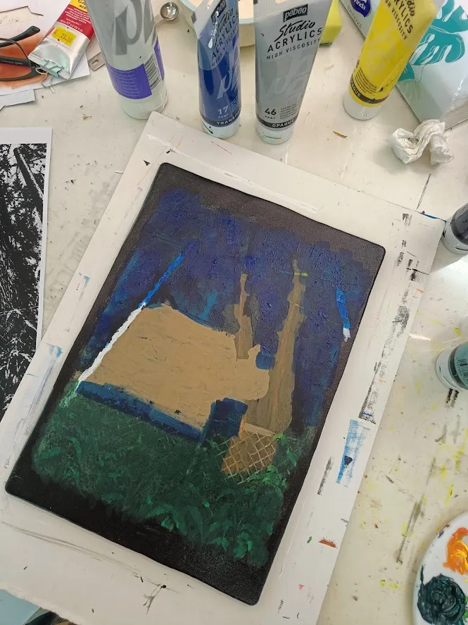 A gel plate in progress on the work table: layers of dark blue, beige and green acrylic paint applied around masks, surrounded by tubes of acrylic paint