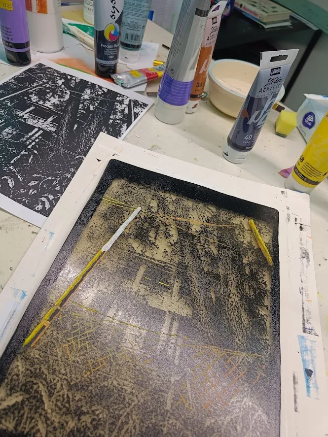 A gel plate in progress on the work table: a dark image transfer of a house among trees, with the printout of the image beside it and tubes of acrylic paint around it