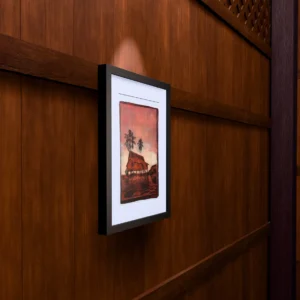 In Pink Sunset, gel plate monotype by Daniel Arendzen, framed on a wooden wall under a spotlight, seen from the side, with binoculars, a watch and a lighter on a table below. The room is a Blender render of the interior of the same traditional Malaysian house shown in the print.