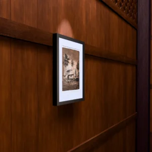 Vintage in Sepia, gel plate monotype by Daniel Arendzen, framed on a wooden wall under a spotlight, seen from the side, with binoculars, a watch and a lighter on a table below. The room is a Blender render of the interior of the same traditional Malaysian house shown in the print.